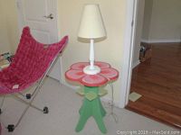 Flower-shaped side table with pink top and green leafy legs next to a red chair, in a room with hardwood floor and yellow walls.