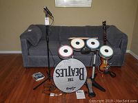 Full view of The Beatles Rock Band electronic drum set with four colored pads, foot pedal, a guitar controller, microphone stand, drum sticks, game discs, and instruction manual arranged in front of a couch.