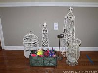 Full view of all decorative metal bird cages of various heights including the tallest which is 30 inches, metal bird figurine on stand, and green metal box containing beaded faux fruit on wooden floor against grey wall.