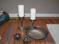 Two tall silver plated candle holders with white pillar candles, small silver plated basket and two small silver plated decorative items on wood floor.