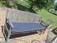 Front view of the larger teak bench showing blue cushion and slatted backrest, placed on outdoor patio.