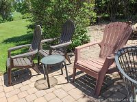 Three resin Adirondack chairs and round outdoor side table arranged on patio with brick pavers and green bushes behind.