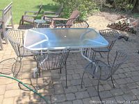 Six metal outdoor chairs with rounded backs around a glass top metal table sitting on a stone paved patio floor. Chairs show rust and wear from outdoor use.