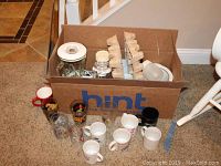 Photo showing a cardboard box filled with a Rival crockpot, a Hamilton blender pitcher, and various mugs and glasses lined in front of the box.