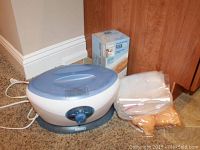 Photo showing the HoMedics paraffin wax machine with wax box and liners placed on carpeted floor near a door and baseboard.