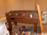 Foosball table in room with other household items. Clear view of top and sides showing players, rods with wooden handles, and wood grain finish.