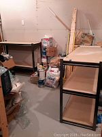 Two identical workbenches in basement space surrounded by boxes and paint supplies. Each has a wood top and wood bottom shelf with black metal framing.