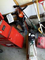 Red toolbox and various garden tools including the electric trimmers and chain saw, photographed in a garage.