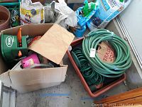 Photo showing a box with a green Scotts hand spreader with orange handles, a pink bottle, garden gloves, and bags of gardening supplies nearby. Next to the box is a rectangular brown self-watering planter tray containing coiled green garden hoses and other garden tools.