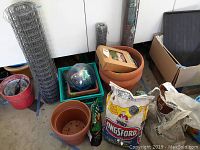 Photo showing two large rolls of chicken wire mesh, plastic planting pots, bags of charcoal and potting soil, gazing ball inside a green crate, and a cardboard box containing garden pebbles.