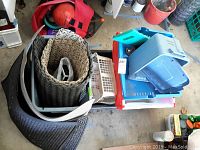 Photo showing a collection of plastic storage containers stacked together with floor door mats arranged nearby in a garage setting.