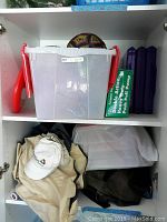 Sports balls, tennis racquets, bleacher pads stored on shelves with plastic containers and baseball caps in garage cabinet.