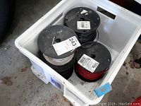 Overhead photo of five rolls of electrical wire in a white plastic bin on a garage floor. Three spools are visible with wire colors black, white, and red.