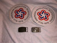 Two round bicentennial decorative platters and two metal belt buckles with bicentennial star emblems, laid on a fabric background.