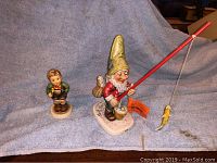 Two vintage Hummel figurines on a blue cloth background. Left is the Bugle Boy holding a bugle, right is Petri fisherman holding a fishing pole with a fish.