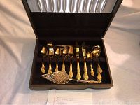 Open flatware set displayed inside fitted black velvet case showing gold-colored spoons, forks, knives, and serving utensils arranged neatly.