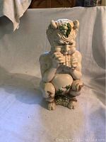 Ceramic figure facing front showing the beast man playing a flute with green clustered accents on head and knees.