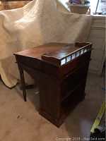 Side angle view of vintage midcentury wooden slant top desk with raised book rail and integrated bookcase below. Shows wooden turned leg, scratches and wear on surfaces, and construction details.