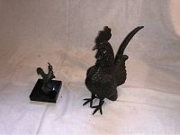 Two cast iron roosters side by side with smaller one on marble base and larger one standing alone on a white cloth background.