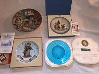 Four collectible plates laid out on fabric, including two Hummel plates in boxes, a blue presidential James Madison plate in original foam casing, and a white Portugal plate with embossed presidential seal.