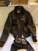 Photo showing full view of worn brown leather bomber jacket with shearling collar and pilot wings pin, alongside vintage leather aviation cap and flight goggles resting below.