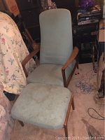 View of mid century Danish teak wood chair with light blue upholstery showing wear and stains, paired with matching ottoman, photographed in indoor setting.