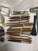 Full view of lot items including pens, belt buckles, pipe, knife, pins, and coins on white background.