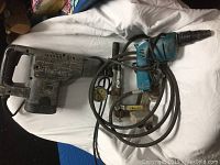 Photo showing two power tools - a large rotary hammer drill and a smaller blue electric drill with cords and several accessories laid out.