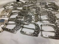 Group of new metal belt buckles with various floral engraved decorations, placed on a white cloth.