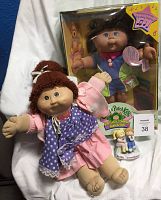 Lot showing two original Cabbage Patch Kids fabric dolls, one dressed in purple and pink outfit, and a boxed doll in blue outfit, with small porcelain figure