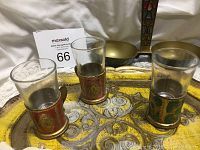 Close-up showing three shot glasses each in colored holders with gold decorative pattern on a vintage metal tray with yellow and gold detailing and floral patterns.