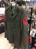 Front and angled view of an olive green military jacket with USMC sergeant chevrons on sleeves and service stripes, buttoned pockets, and epaulets shown on a hanger.