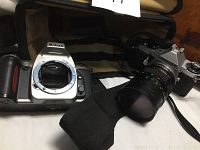 Two cameras and one lens laid on a white surface: Nikon film camera body without lens, Pentax film camera with lens attached and black strap visible