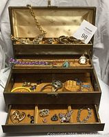 Full view of vintage jewelry box open showing multiple compartments filled with assorted costume jewelry items including bracelets, brooches, earrings, and a watch.