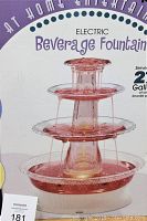 Electric multi-tiered beverage fountain shown inside original packaging, cascading pinkish-red liquid across three transparent tiers.