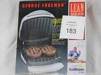 Front view of George Foreman Grill in its original box, showing two burgers on the grill and branding.