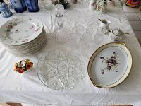 Photo showing collection of 11 floral patterned dinner plates, pressed glass platter, pressed glass decanter with stopper, and six small aperitif glasses arranged on table with floral ceramics.