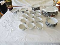 Wide shot showing arranged glassware including wine glasses and brandy snifters, coffee cups, saucers, and plates arranged in stacks; white china with silver rims and floral details