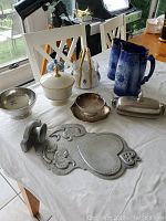 Photo showing multiple items on a white cloth: pewter bowl, Lenox candy bowl with lid, Wedgwood jar with lid, numbered china basket, two blue vintage pitchers, pewter wall sconce and small covered dish.