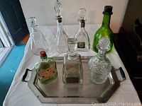 Top view showing all seven vintage glass liquor bottles on the chrome plated brass serving tray with black handles, glass bottles vary in shape and size, some clear and one green bottle visible