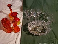 Photo showing red to amber glass decanter, vase, oil and vinegar cruet set, and candy dish on white cloth alongside clear cut glass basket and serving bowl on green cloth.