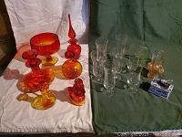 Collection of red to yellow/amber glassware, clear stemware parfait glasses, and ceramic box arranged on green and white fabric backgrounds