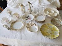 Vintage and antique ceramics and glass collection spread on white cloth tablecloth, includes teapots, plates, cups, saucers, and pitchers, displaying varied designs and gold accents.