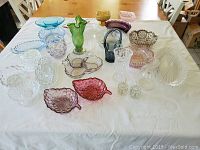 Full view of the collection of various glassware items including pressed glass dishes and vases laid on a white surface under natural light on a wooden table.