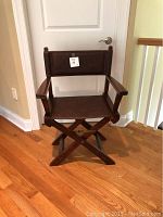 Front view of brown director style folding chair with wooden frame and woven seat and backrest, placed on wooden floor against a closed white door.
