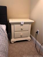 Front and side view of white end table next to a bed, showing two drawers with round knobs and bun feet.