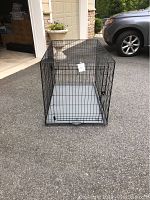 Full view photo of black metal dog crate with removable gray plastic tray inside, positioned outside in a driveway area.