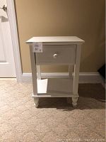 Front view of white nightstand showing drawer and lower shelf standing on carpet.