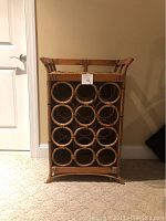 Front view of the rattan and wood wine rack showing 12 circular bottle compartments and woven rattan top, placed against wall with some visible wear.