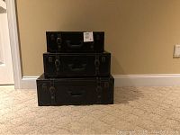 Three black faux leather suitcases stacked largest to smallest on carpet.